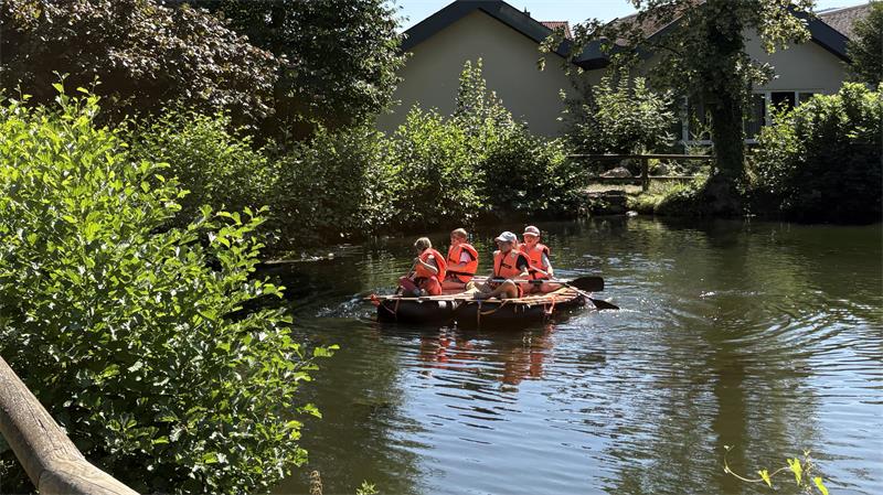 Gruppe von Kindern mit roten Schwimmwesten auf einem Floß auf einem ruhigen Fluss vor einem Haus und umgeben von Bäumen