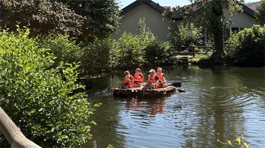 Gruppe von Kindern mit roten Schwimmwesten auf einem Floß auf einem ruhigen Fluss vor einem Haus und umgeben von Bäumen