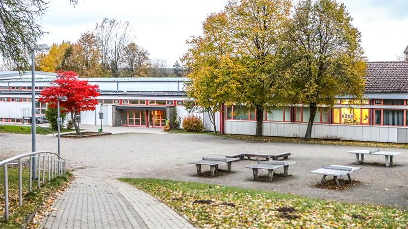 Schulhof mit Bänken und Tischen, umgeben von Bäumen mit herbstlich gefärbtem Laub und einem Schulgebäude im Hintergrund
