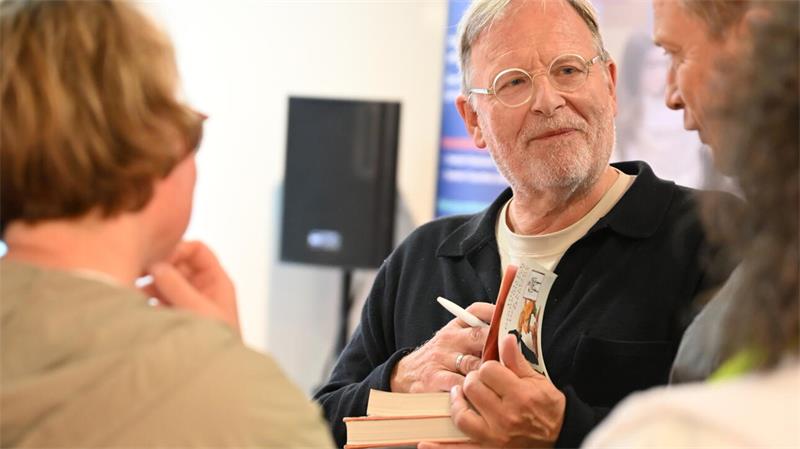 Älterer Mann mit Brille signiert ein Buch und unterhält sich mit mehreren Personen in einem Innenraum