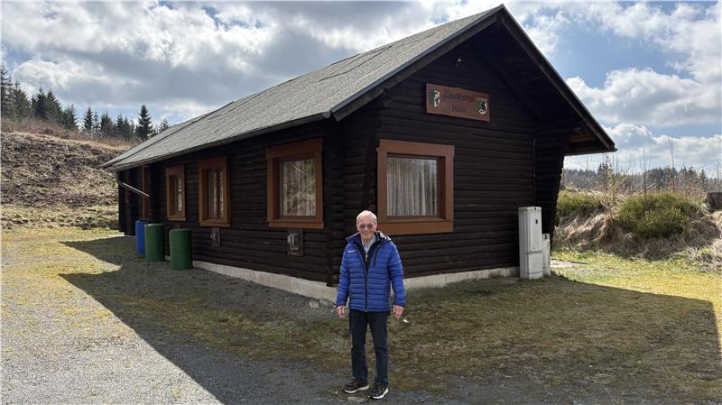 Mann in blauer Jacke steht vor einem dunkelbraunen Holzhaus mit mehreren Fenstern und einem Schild über der Tür