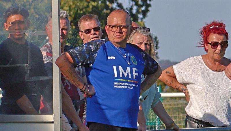 Mehrere Personen stehen hinter einem Metallgeländer, eine Person trägt ein blaues Trikot mit der Aufschrift 'IMF' und hat die Hände in die Hüften gestützt