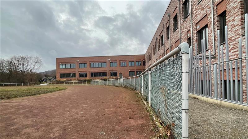 Blick entlang eines Maschendrahtzauns auf einen roten Backsteinbau und ein modernes Gebäude unter bewölktem Himmel