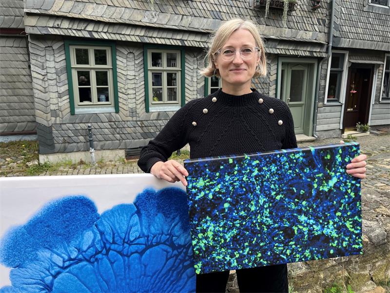 Eine blonde Frau mit Brille steht vor einem grauen Haus und hält zwei blaue Gemälde in den Händen.