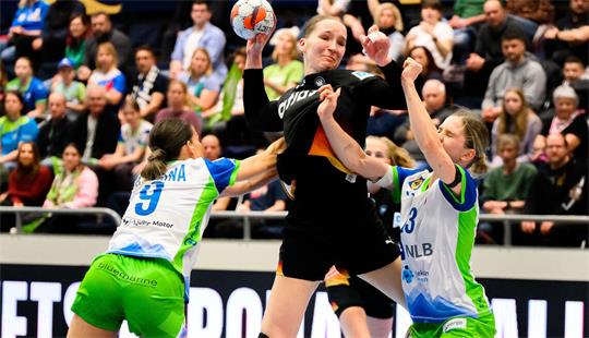 Drei Handballspielerinnen im Sprung, zwei in weißen Trikots mit grünen Hosen verteidigen gegen eine Spielerin in schwarzem Trikot mit Ball in der Hand vor Zuschauern auf der Tribüne