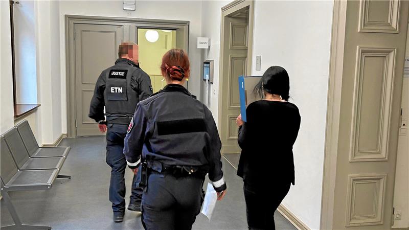 Zwei Justizbeamte und eine Frau, die sich einen Ordner vors Gesicht hält, gehen über einen Flur.