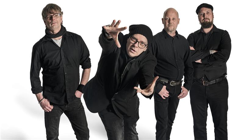 Vier Männer stehen in schwarzen Klamotten und posen vor weißem Hintergrund. 