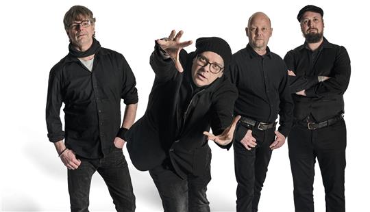 Vier Männer stehen in schwarzen Klamotten und posen vor weißem Hintergrund. 