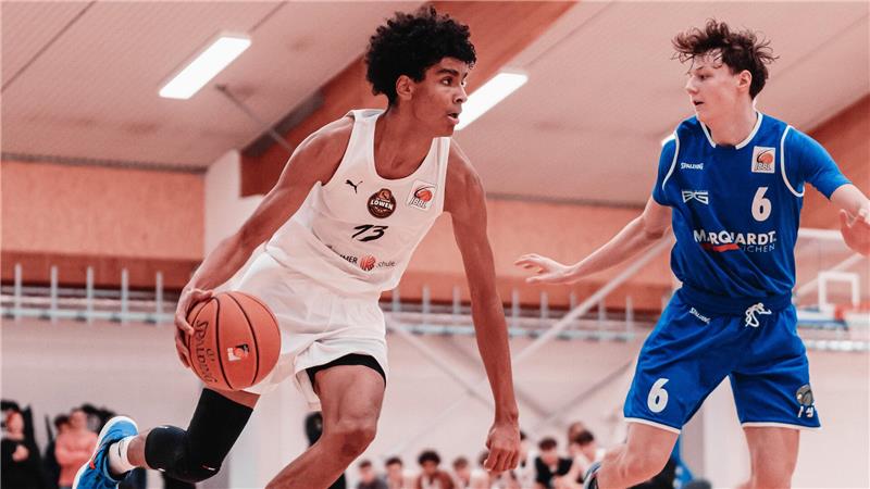 Zwei Basketballspieler in Aktion, einer in weißem Trikot mit Ball, der andere in blauem Trikot verteidigt.