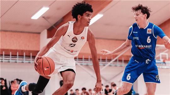 Zwei Basketballspieler in Aktion, einer in weißem Trikot mit Ball, der andere in blauem Trikot verteidigt.