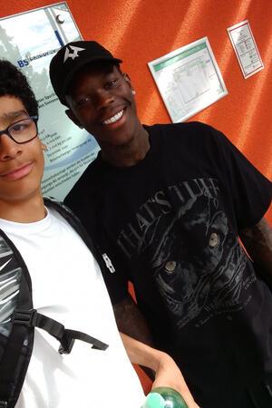 Ein Goslarer auf den Spuren von Dennis Schröder Zwei Personen vor orangefarbener Wand mit mehreren Aushängen, eine trägt schwarzen Pullover mit Tigerkopf und schwarze Kappe mit weißem Stern, die andere weißen T-Shirt und schwarzen Rucksack.