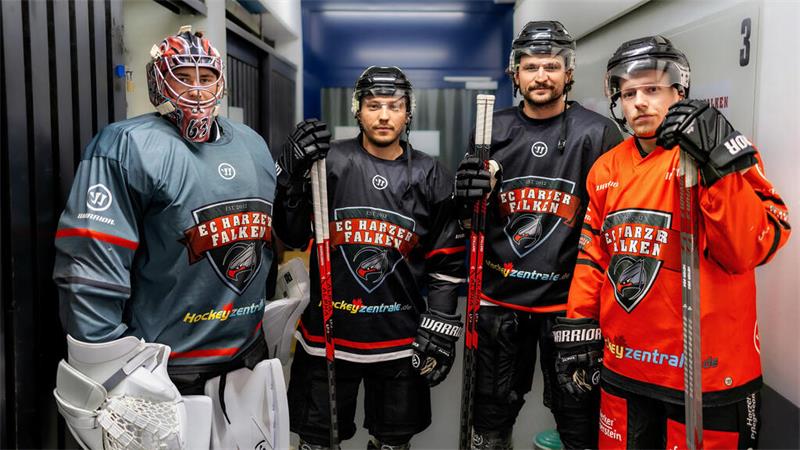 Vier Eishockeyspieler in voller Ausrüstung stehen in einem Flur, drei tragen schwarze Trikots mit dem Schriftzug 'EC Harzer Falken', einer ein rotes Trikot, alle mit Helm und Schläger