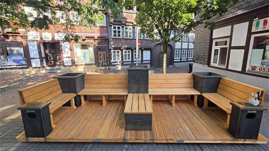 Holzterrasse mit L-förmigen Sitzbänken, quadratischen Pflanzkästen und einem kleinen Tisch in einem Innenhof mit Fachwerkhäusern im Hintergrund