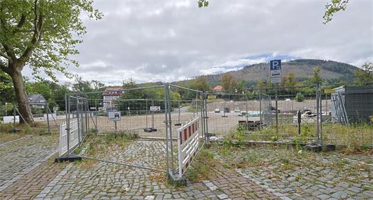 Offener Bauzaun auf gepflastertem Platz mit Bäumen und Hügeln im Hintergrund