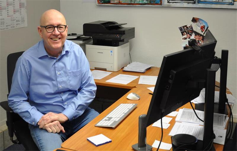 Mann mit blauem Hemd sitzt an einem Schreibtisch mit Computer, Tastatur, Maus und mehreren Papierstapeln in einem Büro