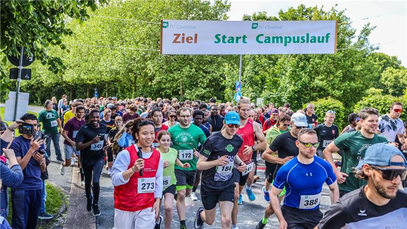 Läufer starten bei einem Campuslauf unter einem Banner mit der Aufschrift 'Ziel Start Campuslauf' auf einem von Bäumen gesäumten Weg.