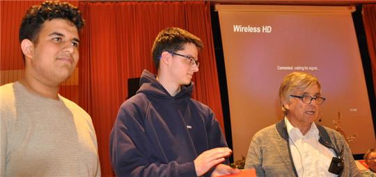 Drei Personen stehen vor einem roten Vorhang und einem Projektorbildschirm mit der Anzeige 'Wireless HD Connected, waiting for signal'