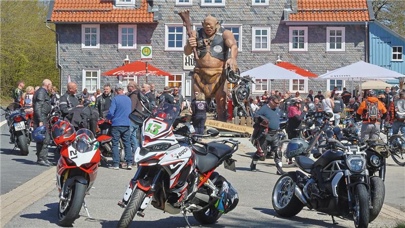 Vor einem Haus steht ein Riese als Skulptur, darum herum sitzen und stehen Menschen unter Sonnenschirmen, im Vordergrund einige Motorräder.