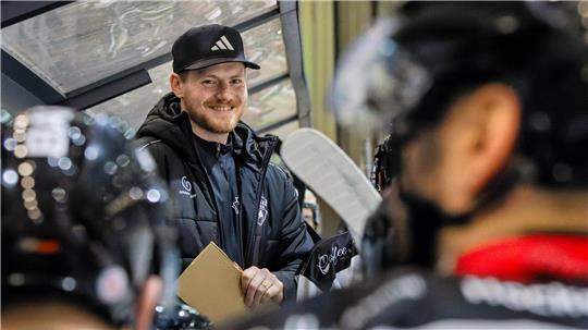 Mann mit schwarzer Adidas-Kappe und schwarzer Jacke hält ein Klemmbrett und steht hinter einer Plexiglasscheibe vor mehreren Eishockeyspielern mit Helmen.