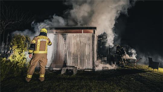 Feuerwehrmann in Schutzkleidung vor einem kleinen brennenden Holzschuppen, aus dem Rauch aufsteigt, nachts im Garten.