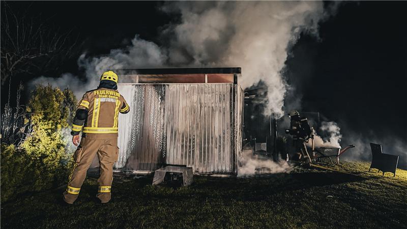 Feuerwehrmann in Schutzkleidung vor einem kleinen brennenden Holzschuppen, aus dem Rauch aufsteigt, nachts im Garten.