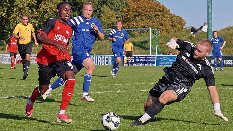 Fußballspieler in Aktion auf dem Spielfeld, ein Spieler in rotem Trikot läuft am Ball vorbei, Torwart in schwarzem Trikot rutscht zum Ball