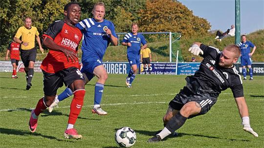 Fußballspieler in Aktion auf dem Spielfeld, ein Spieler in rotem Trikot läuft am Ball vorbei, Torwart in schwarzem Trikot rutscht zum Ball