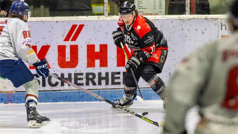 Eishockeyspieler in schwarzer und roter Ausrüstung mit Puck vor zwei Gegnern auf dem Eis