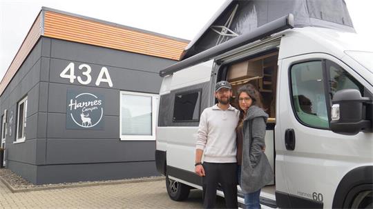 Ein Mann und eine Frau stehen vor einem weißen Wohnmobil mit ausgefahrenem Aufstelldach. Im Hintergrund befindet sich das dunkelgraue Gebäude eines Hannes-Camper-Standorts mit der Hausnummer 43A und dem Firmenlogo.