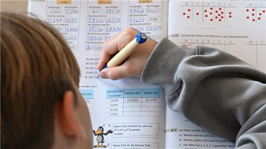 Kind mit braunen Haaren schreibt mit blauem Stift auf einem Arbeitsblatt mit Zahlen und Tabellen