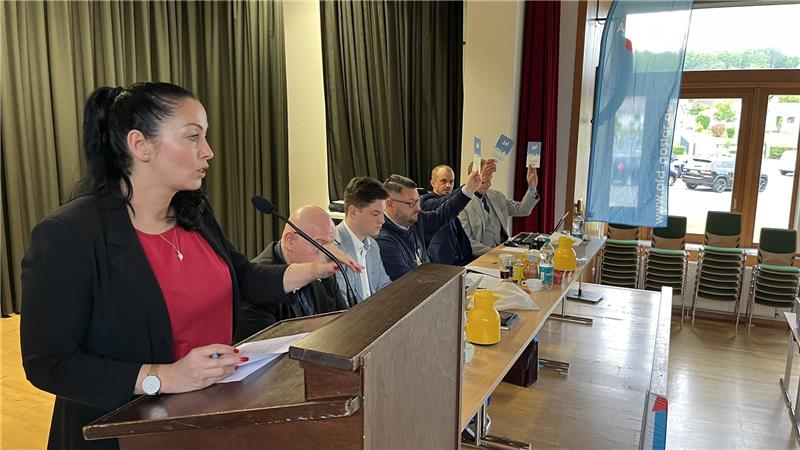 Eine Frau steht an einem Pult und spricht. Neben ihr sitzen fünf Männer.