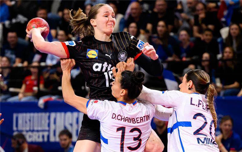 Handballspiel: Spielerin in schwarzem Trikot springt mit Ball, zwei Spielerinnen in weißen Trikots verteidigen mit erhobenen Armen.