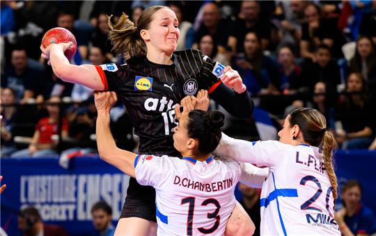 Handballspiel: Spielerin in schwarzem Trikot springt mit Ball, zwei Spielerinnen in weißen Trikots verteidigen mit erhobenen Armen.