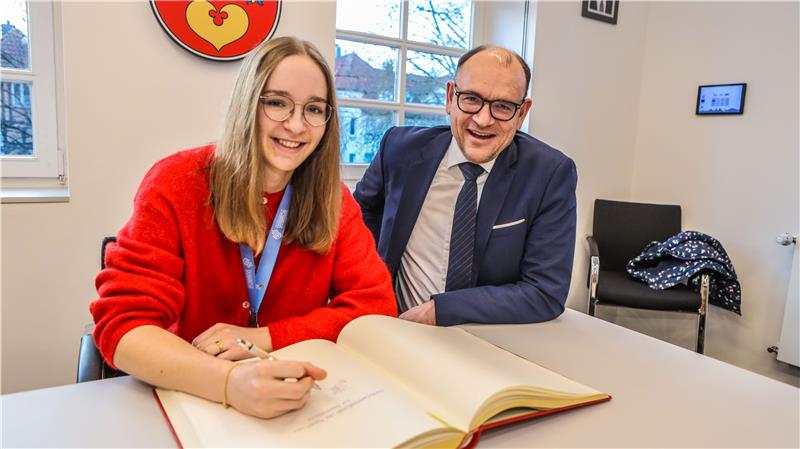 Zwei Personen sitzen an einem Tisch, eine trägt einen roten Pullover und schreibt in ein aufgeschlagenes Buch, die andere trägt einen Anzug und Krawatte.