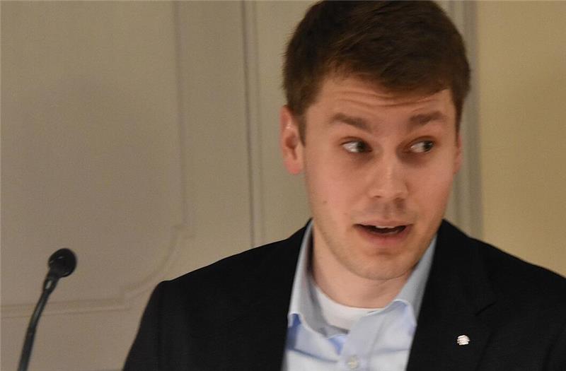 Mann mit dunklem Sakko und hellblauem Hemd vor einer Wand mit Mikrofonständer