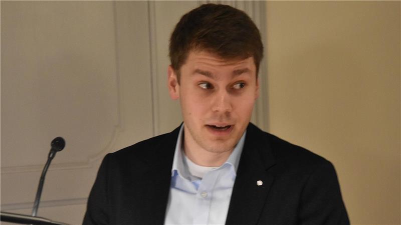 Mann mit dunklem Sakko und hellblauem Hemd vor grauer Wand stehend mit Mikrofon im Vordergrund