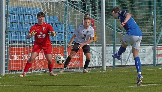 Fußballspieler in blauer Trikotfarbe schießt auf das Tor, Torwart und Verteidiger in roten und weißen Trikots stehen im Torraum bereit