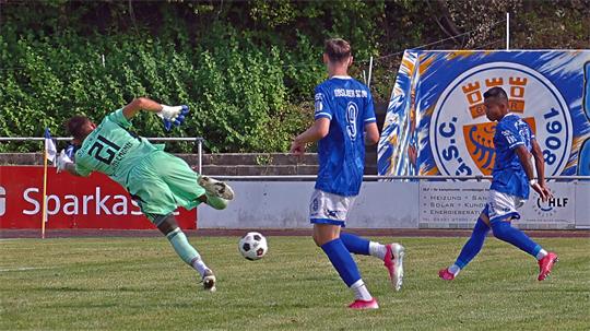 Torwart in grün springt nach links, zwei Spieler in blauen Trikots beobachten den Ball auf dem Spielfeld