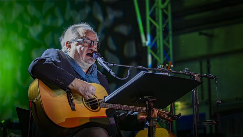 Hein Rudolf Kunze sitzt auf der Bühne mit Gitarre und singt ins Mikrofon.