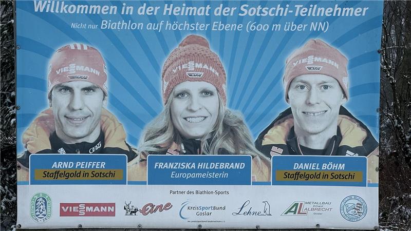 Banner mit drei Biathleten in Sportkleidung und Mützen vor blauem Hintergrund mit Strahlenmuster und Text "Willkommen in der Heimat der Sotschi-Teilnehmer" sowie deren Namen und Auszeichnungen.