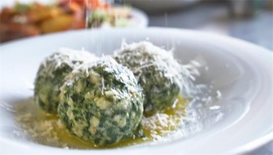 Auf einem Teller liegen Spinatknödel, jemand streut Käse darüber.