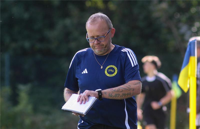 Mann in blauem Sportshirt mit gelbem Logo hält Notizblock auf einem Fußballfeld mit Eckfahne und weiteren Personen im Hintergrund