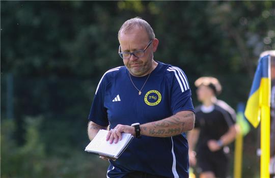 Mann in blauem Sportshirt mit gelbem Logo hält Notizblock auf einem Fußballfeld mit Eckfahne und weiteren Personen im Hintergrund