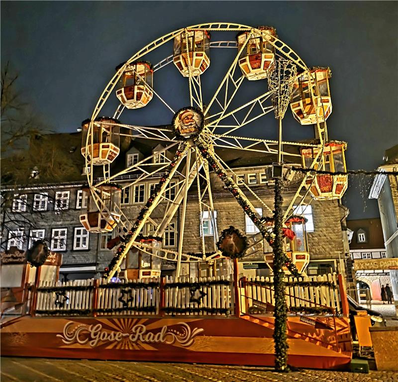 Beleuchtetes Riesenrad mit geschlossenen Gondeln auf einem Platz vor Fachwerkhäusern bei Nacht.