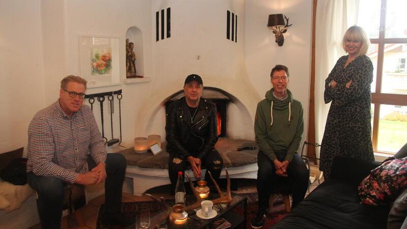 Das Foto zeigt drei Männer, die vor einem brennenden Kamin sitzen und eine Frau, die am Fenster daneben steht.