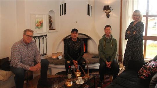 Das Foto zeigt drei Männer, die vor einem brennenden Kamin sitzen und eine Frau, die am Fenster daneben steht.