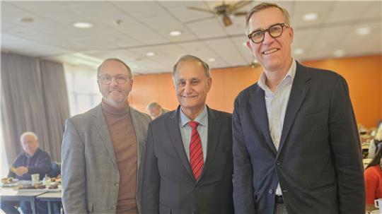 Drei Männer in Anzügen stehen in einem Raum nebeneinander.