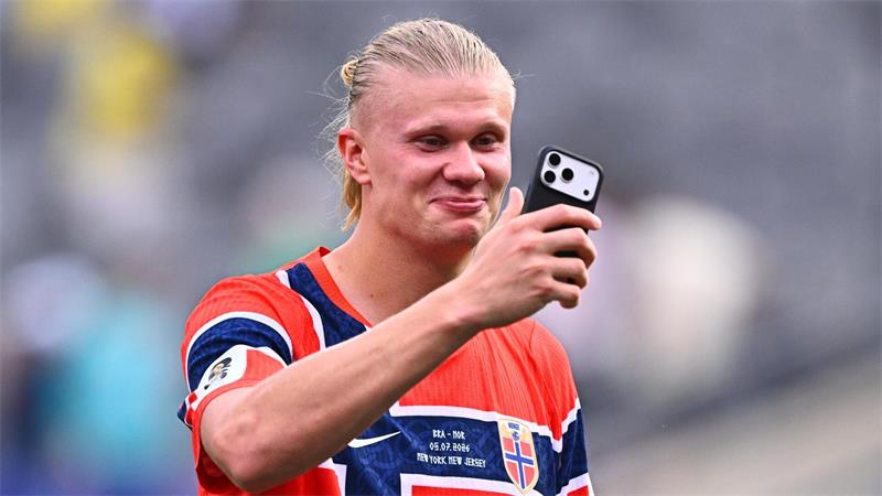 Junger Mann mit blondem Zopf trägt ein norwegisches Fußballtrikot und hält ein Smartphone in der Hand