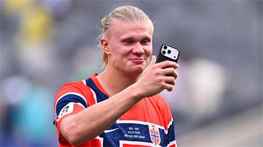 Junger Mann mit blondem Zopf trägt ein norwegisches Fußballtrikot und hält ein Smartphone in der Hand