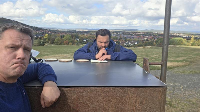 Zwei Männer sitzen an einem Tisch im Freien mit Blick auf eine Landschaft und einen Ort im Hintergrund